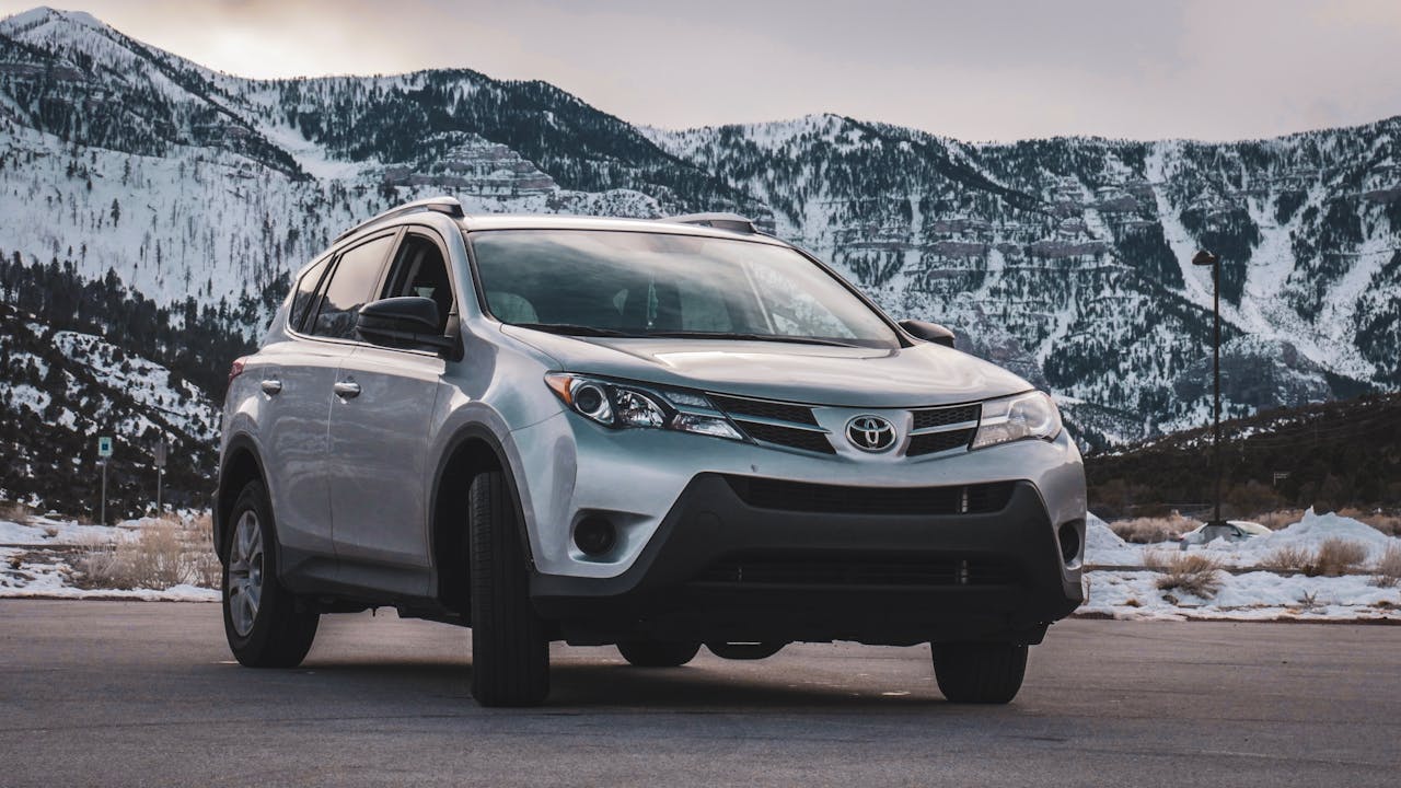 A silver SUV is parked in front of stunning snow-covered mountains during daytime.